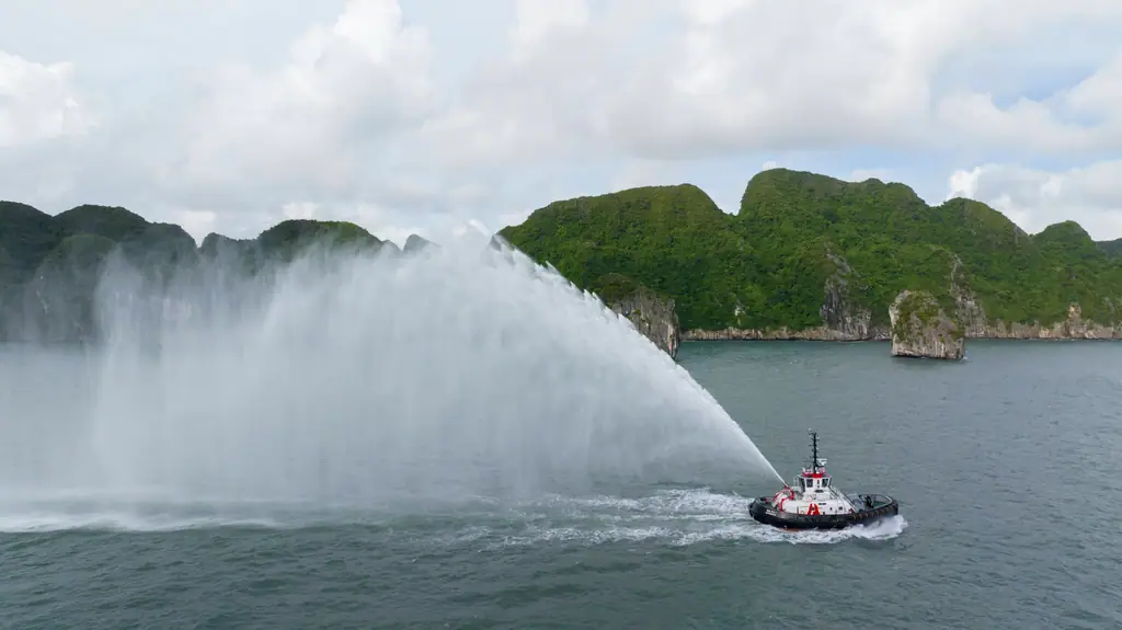 Tug with its fire monitors running, seen from the air, in a bay with green islands