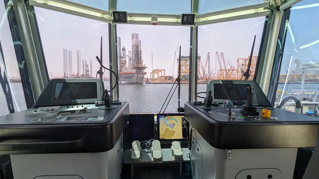 View from a wheelhouse over the water, with jack-up rigs and cranes at a yard on the far side